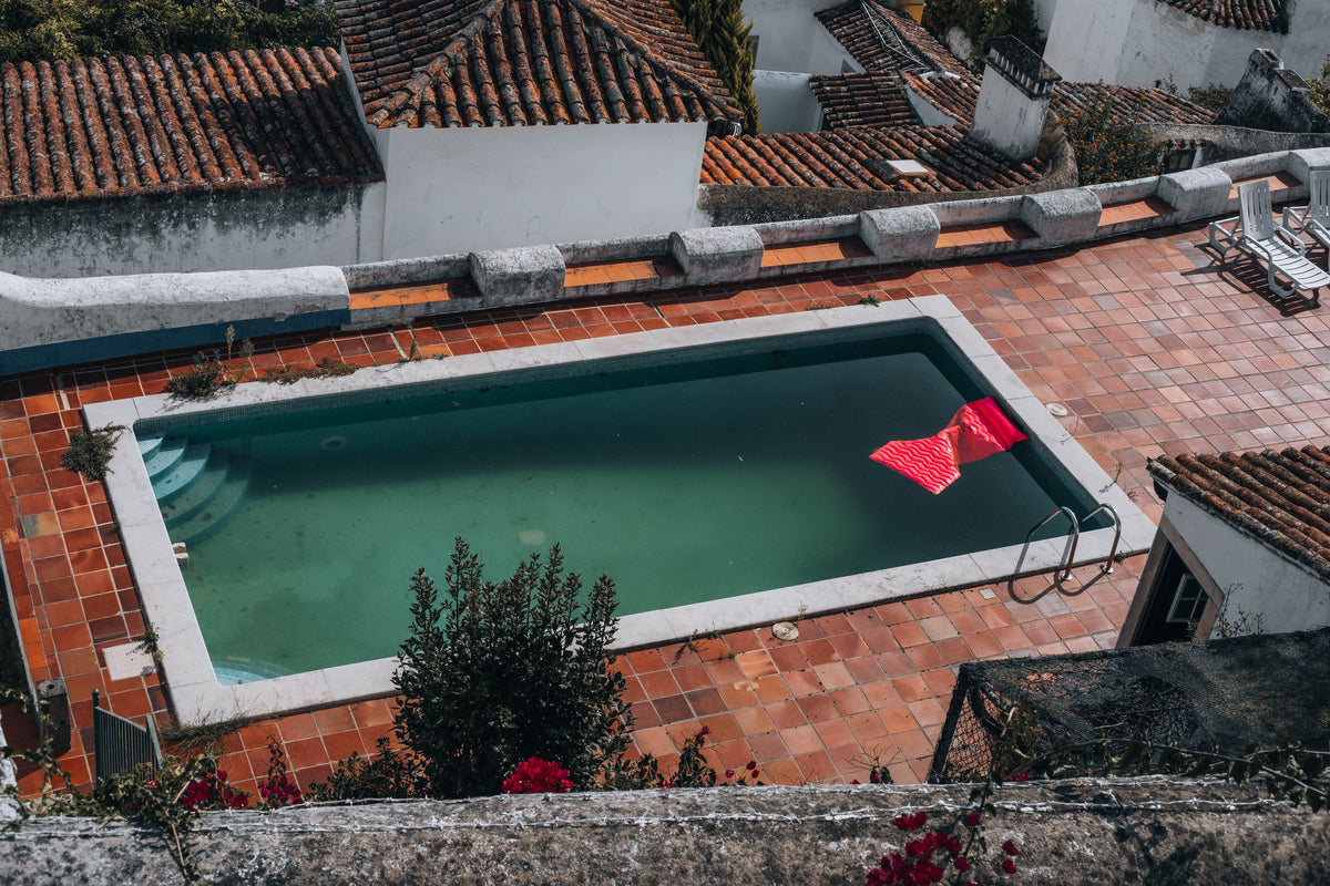 An aerial view of a rectangular rooftop swimming pool with terracotta tile decking, white walls, and red-tiled roofs in the background. A red patterned float is floating on the pool water.